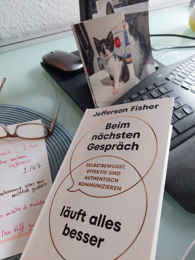 Buch „Beim nächsten Gespräch läuft alles besser“ von Jefferson Fisher liegt auf einem Schreibtisch neben einer Brille, handschriftlichen Notizen und einer Katzenbild.